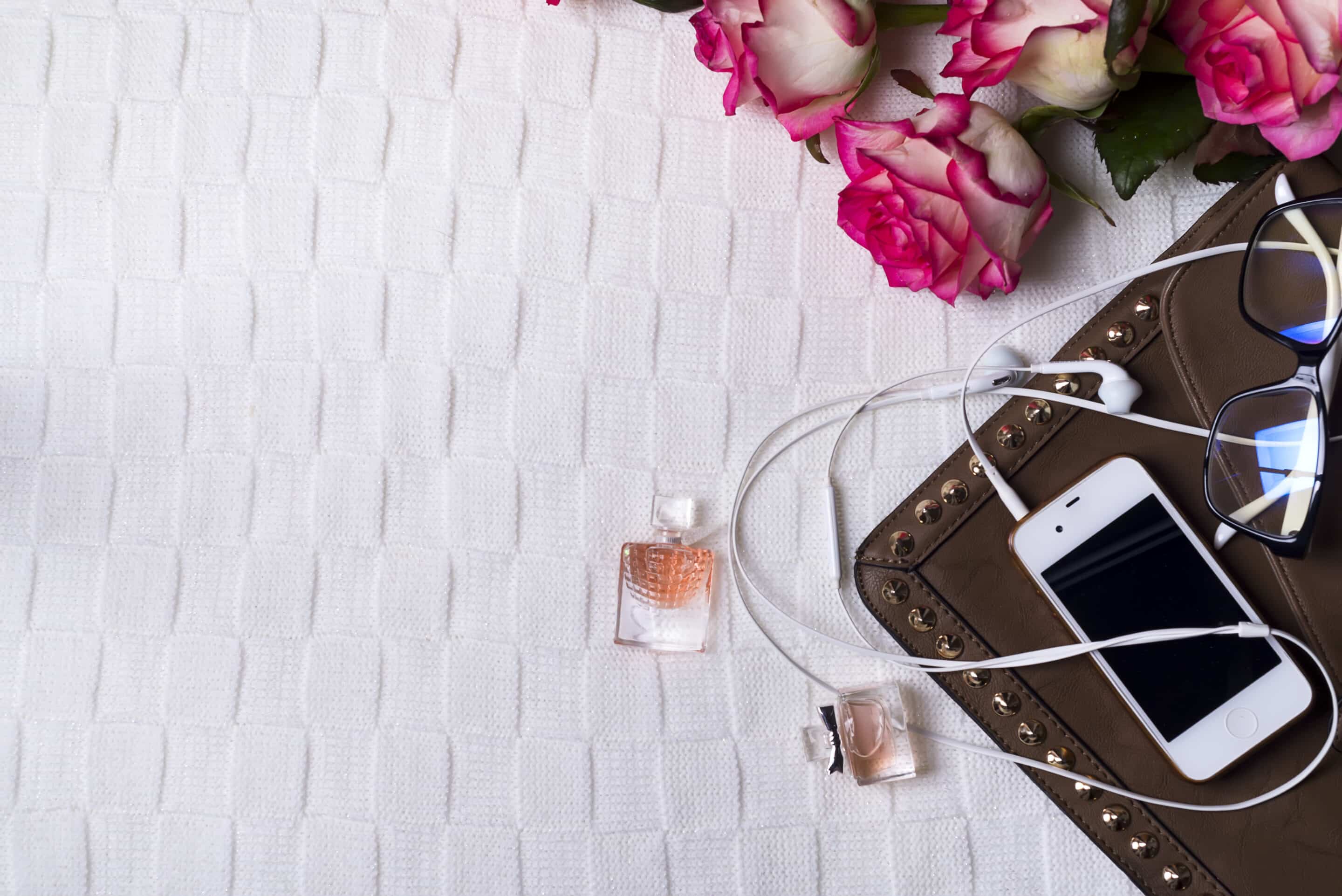 female clutch with phone, headphones and Perfume bottles with flowers on light background. Perfumery, cosmetics, fragrance collection.